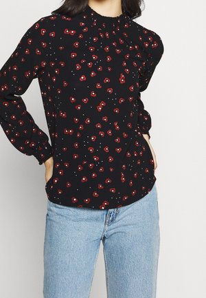 Blusa negra con estampado floral rojo, con escote fruncido y mangas largas abullonadas. Combinada con unos pantalones de mezclilla azul claro.