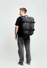 Mochila negra impermeable con un gran logo, múltiples correas ajustables, un asa resistente y cremalleras; de forma rectangular con una textura lisa.
