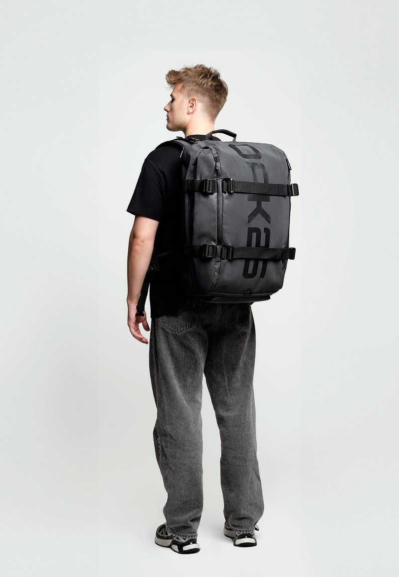 Mochila negra impermeable con un gran logo, múltiples correas ajustables, un asa resistente y cremalleras; de forma rectangular con una textura lisa.