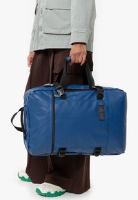 Blå duffelväska gjord av hållbart material, med svarta detaljer, topphandtag och dragkedjestängning, hålls av en person som bär bruna byxor.