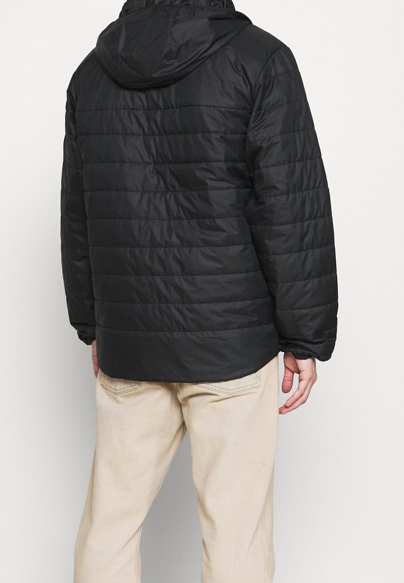 Svart vadderad jacka med huva, med horisontellt quiltat mönster och elastiska ärmslut. Bärs över ljusbeiga byxor.