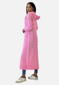 Długi różowy sweter kardigan z kapturem, ozdobiony wzorem w warkocze i ściągaczami na mankietach. W połączeniu z jasnoniebieskimi dżinsami i białymi trampkami.