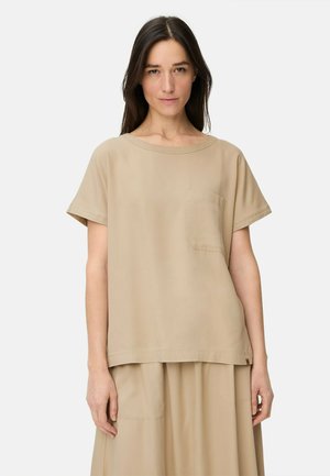 Vrouw met lang donker haar die een beige top met korte mouwen en borstzakje draagt, en een bijpassende beige rok, staand tegen een witte achtergrond.