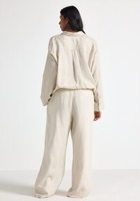 Beige lněný outfit obsahující volnou košili s plisovaným zadním dílem a širokými kalhotami, zdůrazňující jemnou strukturu a neutrální barvu.