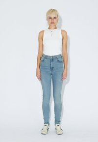 Persona con pelo rubio corto que lleva una camiseta sin mangas blanca, unos jeans de tiro alto en azul claro, zapatillas blancas y collares en capas, de pie.