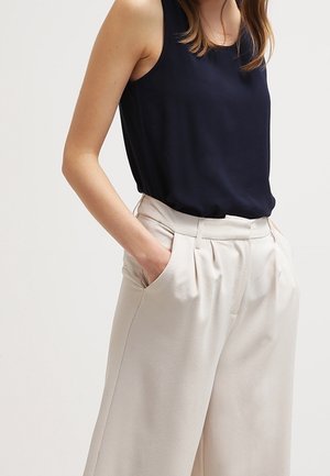 Femme portant un chemisier noir sans manches, rentré dans un pantalon beige taille haute à jambes larges, les mains dans les poches.