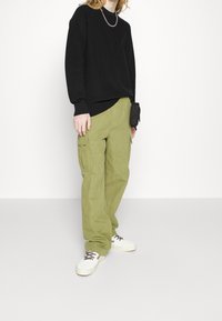 Zwarte sweatshirt met geribde manchetten, gecombineerd met olijfgroene cargo pants met zijzakken en een zwarte tas. Witte sneakers maken de look compleet.