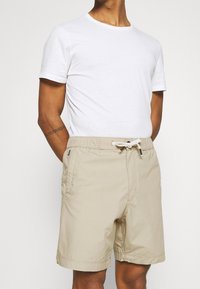Man wearing a plain white short-sleeve t-shirt and beige drawstring shorts, standing with hands in pockets against a plain background.