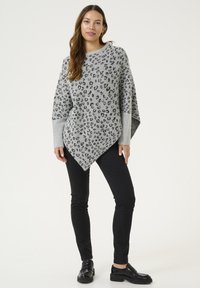 Grauer Pullover mit schwarzem Leopardenmuster, oversized Design mit V-förmigem Saum. Kombiniert mit schwarzen Slim-Fit-Hosen und klobigen schwarzen Schuhen.
