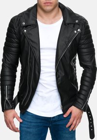 Giacca da biker in pelle nera con spalle trapuntate, cerniere argentate e colletto a pressione. Indossata sopra una camicia bianca e abbinata a jeans blu.