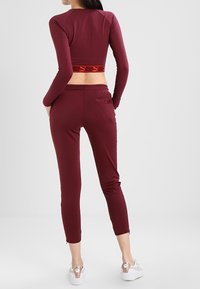 Burgunderfarbenes Sportset bestehend aus einem langen Cropped-Top mit Logo-Band und engen Hosen. Glatte Textur, dehnbare Stoffe und stromlinienförmige Silhouette.
