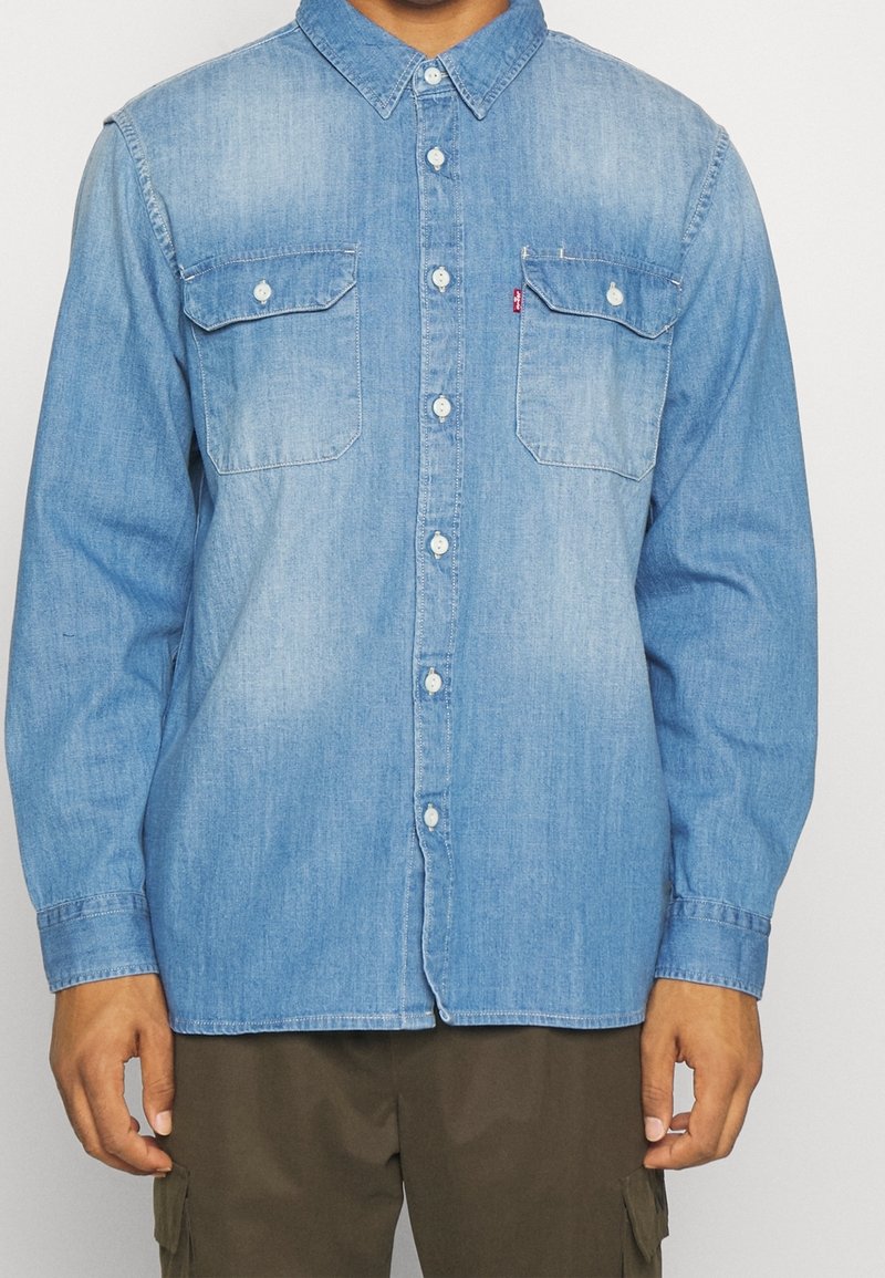 Chemise en denim bleu avec col boutonné, deux poches poitrine et manches longues. Le tissu a une texture lisse avec un léger effet délavé.