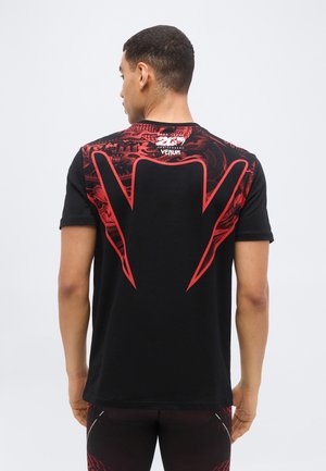 Άνδρας που κοιτάζει προς τα πίσω φοράει μαύρο μπλουζάκι με μεγάλο κόκκινο γωνιακό σχέδιο και διακριτικά γραφικά μοτίβα, συνδυασμένο με μαύρο και κόκκινο παντελόνι.