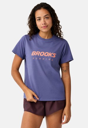 Vrouw met krullend haar die een paarse Brooks Running T-shirt en donkere korte broek draagt, staand met één hand die de zoom van het shirt vasthoudt.