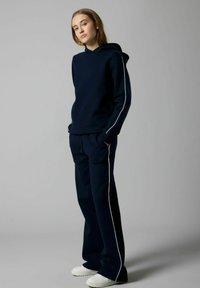 Conjunto de sudadera y pantalones de chándal en color navy con franjas laterales blancas. Material suave, corte relajado y puños acanalados. Zapatillas blancas que completan el conjunto.