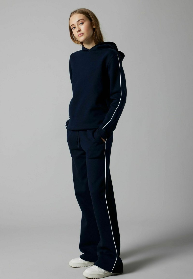 Conjunto de sudadera y pantalones de chándal en color navy con franjas laterales blancas. Material suave, corte relajado y puños acanalados. Zapatillas blancas que completan el conjunto.