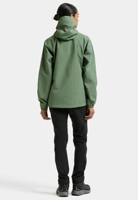 Grüne wasserdichte Jacke mit Kapuze, elastischen Bündchen und lockerem Sitz, kombiniert mit schwarzen Hosen und grauen Wanderschuhen. Rückansicht gezeigt.