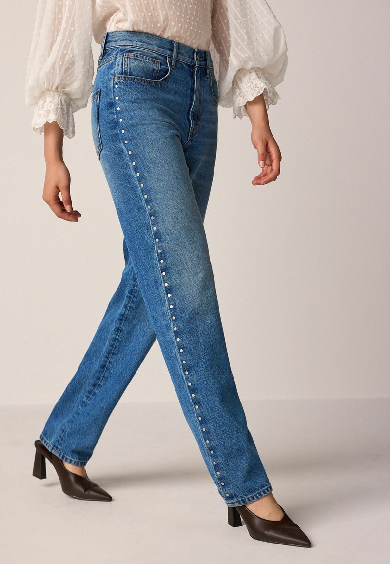 Femme marchant portant un jean bleu taille haute avec des clous argentés sur le côté et des chaussures marron à talons blocs à bout pointu.