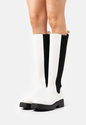 Botas brancas e pretas até ao joelho com um acabamento suave, apresentando uma sola preta robusta e um design de painel vertical em contraste.
