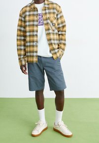 Chemise en flanelle écossaise jaune et noire, t-shirt blanc à motifs en dessous, shorts gris, chaussettes blanches et baskets blanches avec semelles marron.
