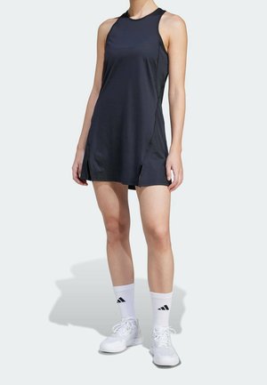 Kobieta ubrana w czarną, sportową, bezrękawnikową sukienkę, białe skarpety Adidas oraz białe buty sportowe, stojąca na jednolitym tle.