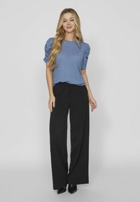 Modrá blúzka s puffovými rukávmi a štruktúrovaným vzorom, kombinovaná s vysokými čiernymi nohavicami s širokými nohavicami a čiernymi lodičkami s ostrou špičkou.