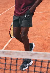 Musta Nike tennis-shortsit, joissa on sivutaskut, harmaat ja valkoiset Nike-kengät, seisomassa savikentällä. Maila osittain näkyvissä.