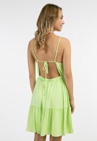 Rochie de vară verde deschis, cu fustă stratificată, bretele subțiri și design cu spate deschis, având un detaliu cu funda la talie pentru ajustare.