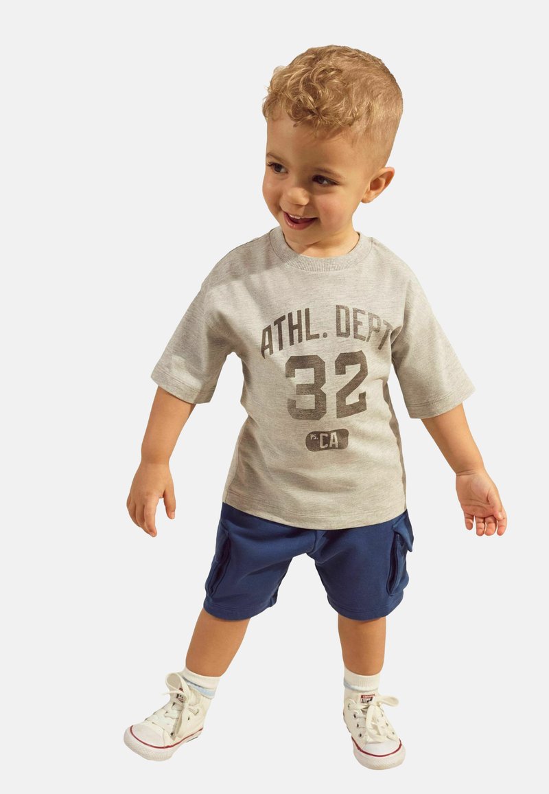 T-shirt gris à manches courtes avec un motif foncé, associé à un short cargo bleu marine et des baskets blanches. Enfant souriant et se tenant dans une pose ludique.