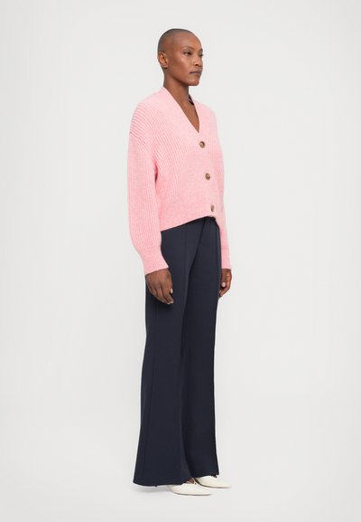 Rosa gerippter Cardigan mit V-Ausschnitt und drei Holzknöpfen, kombiniert mit navyblauen weit geschnittenen Hosen und weißen spitzen Schuhen.