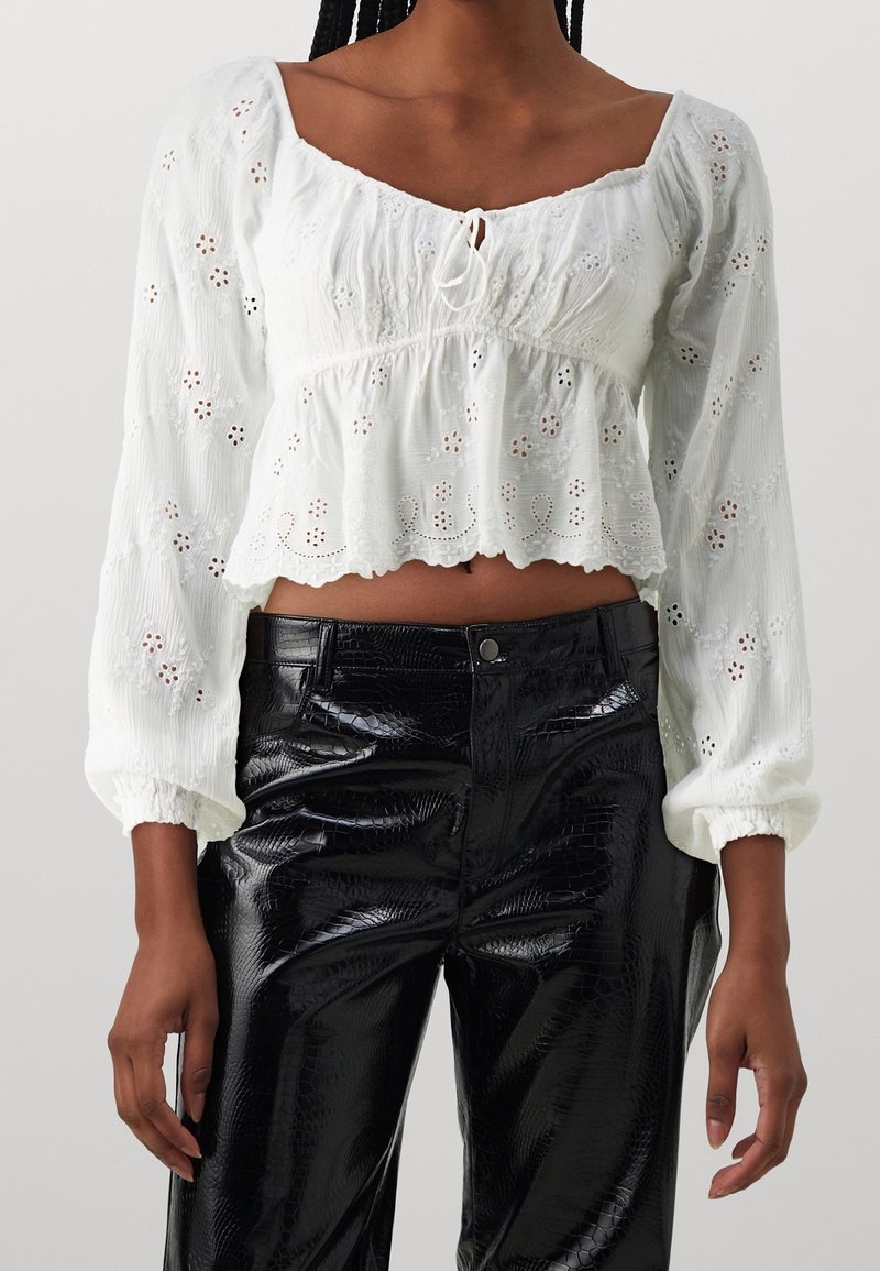 Žena v bílém dlouhém rukávu s krajkovou zkrácenou blůzkou a černých vysokých lesklých kalhotách.