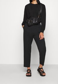 Haut noir à manches longues avec bretelles volantées ornées de clous, pantalon noir taille haute, sandales noires à brides, accompagné d'un petit sac noir.