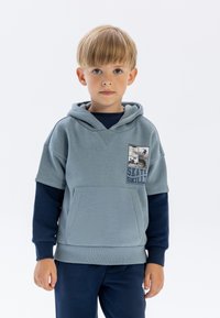 Sudadera capucha azul claro con bolsillo frontal, que presenta un estampado gráfico. Llevada sobre una camiseta de manga larga azul marino, con pantalones a juego en azul marino.