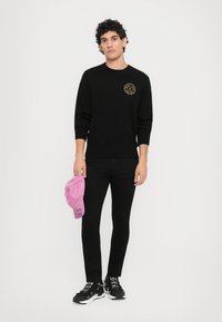 Μαύρη βαμβακερή μπλούζα με στρογγυλή λαιμόκοψη και χρυσό λογότυπο, συνδυασμένη με μαύρα στενά τζιν παντελόνια, κρατώντας ένα ροζ καπέλο με κείμενο. Μαύρα αθλητικά παπούτσια.