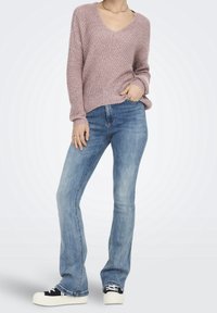 Maglione rosa chiaro lavorato a maglia con scollatura a V profonda, abbinato a jeans svasati blu e scarpe da ginnastica con plateau nero e bianco. Dettagli del tessuto in evidenza.