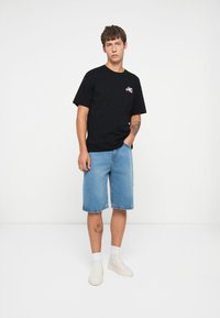 Black cotton t-shirt with front graphic, paired with light blue denim shorts. Model wears white slip-on shoes and white socks.