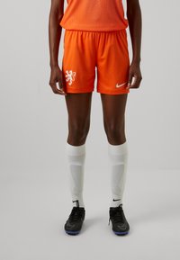 Oranžové futbalové šortky s bielym emblémom leva a logom Nike. V kombinácii s bielymi kolennými ponožkami a čiernymi kopačkami. Hladký, priedušný materiál.