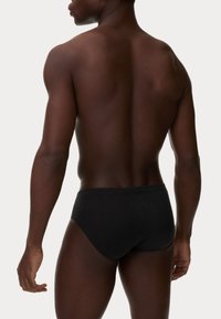 Schwarze Schwimmslips, getragen von einem Modell mit einem glatten, dunklen Hautton. Der Stoff wirkt weich mit einem nahtlosen Design und sitzt tief auf den Hüften.