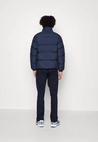 Mörkblå puffjacka med hög krage och horisontell sömnad, som bärs över svarta byxor och mönstrade sneakers, sedd bakifrån.