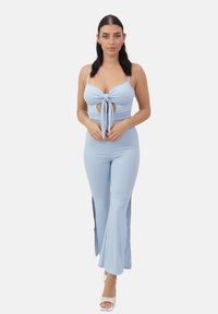 Mono de color azul claro con acanalado que presenta un lazo frontal y detalles cut-out, con piernas acampanadas y aberturas laterales. Llevado con sandalias de tacón blancas.