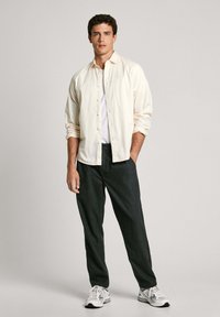 Gai krēmu pogkrekls virs balta T-kreklu, saskaņots ar tumši pelēkiem biksēm un baltām kedām, stāvot uz vienkārša fona.