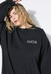 Svart oversized sweatshirt i mjukt material med ordet "AMOUR" i grått, med rund halsringning och avslappnad passform.