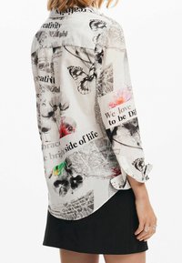 Langsleeve, loszittend overhemd van doorschijnende witte stof met bloemen- en vlinderprints, gemengde tekst elementen en opgerolde mouwen.