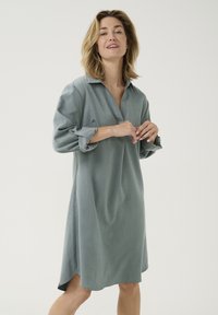 Robe chemise bleu clair avec un col, des manches longues retroussées aux poignets et une fente latérale pour faciliter le mouvement. Tissu doux et texturé.