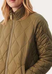 Olivgrüne Steppjacke mit hohem Kragen, diagonaler Naht und goldenen Reißverschlussdetails. Weicher, leichter und strukturierter Stoff.