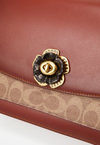 Kopertówka z ciemnoczerwonej skóry, z teksturowanym beżowym panelem materiałowym oraz klamrą w kształcie kwiatu w kolorze czarnym i złotym.