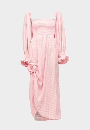 Femme portant une longue robe vichy rose avec des manches bouffantes et un détail de rose en tissu près d'une fente frontale, associée à des sandales slides beiges.