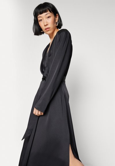 Μαύρο μακρυμάνικο φόρεμα σε φάκελο από απαλό ύφασμα, με βαθύ ντεκολτέ, ζώνη στη μέση και ύψος σκισίματος στο μπουστό. Μινιμαλιστικός σχεδιασμός.