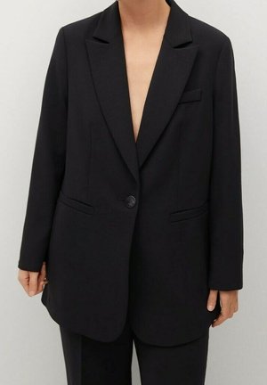 Person iført en sort enkeltknappet blazer med hakkede revers og to forlommer, kombineret med sorte bukser mod en ensfarvet baggrund.