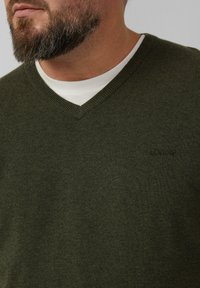 Maglione a V in misto lana verde scuro, con una texture lavorata a maglia e un piccolo logo sul petto, indossato sopra una camicia bianca.